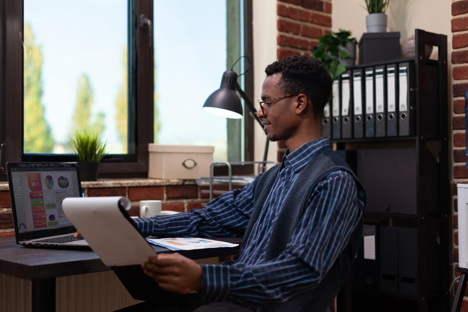 Smiling small business owner looking at laptop screen with analytics data holding clipboard sitting at desk.