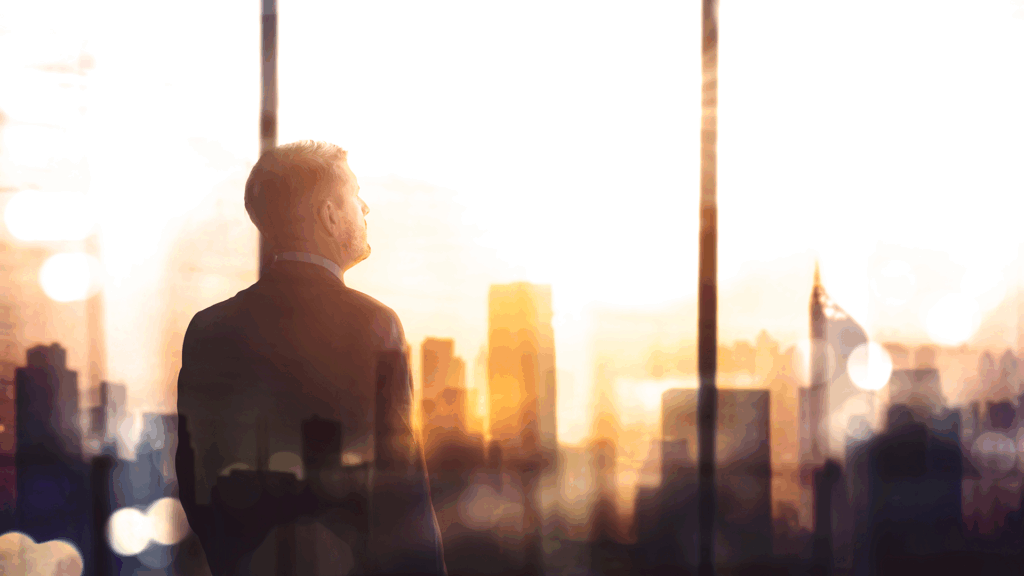 Business professional in a suit looking out over a city skyline from a high-rise office as the sun rises, symbolizing vision and future opportunities.