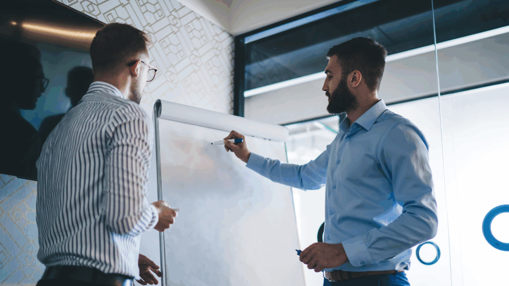 Two professionals collaborating at a whiteboard in a modern office, brainstorming or discussing strategy with marker pens in hand.