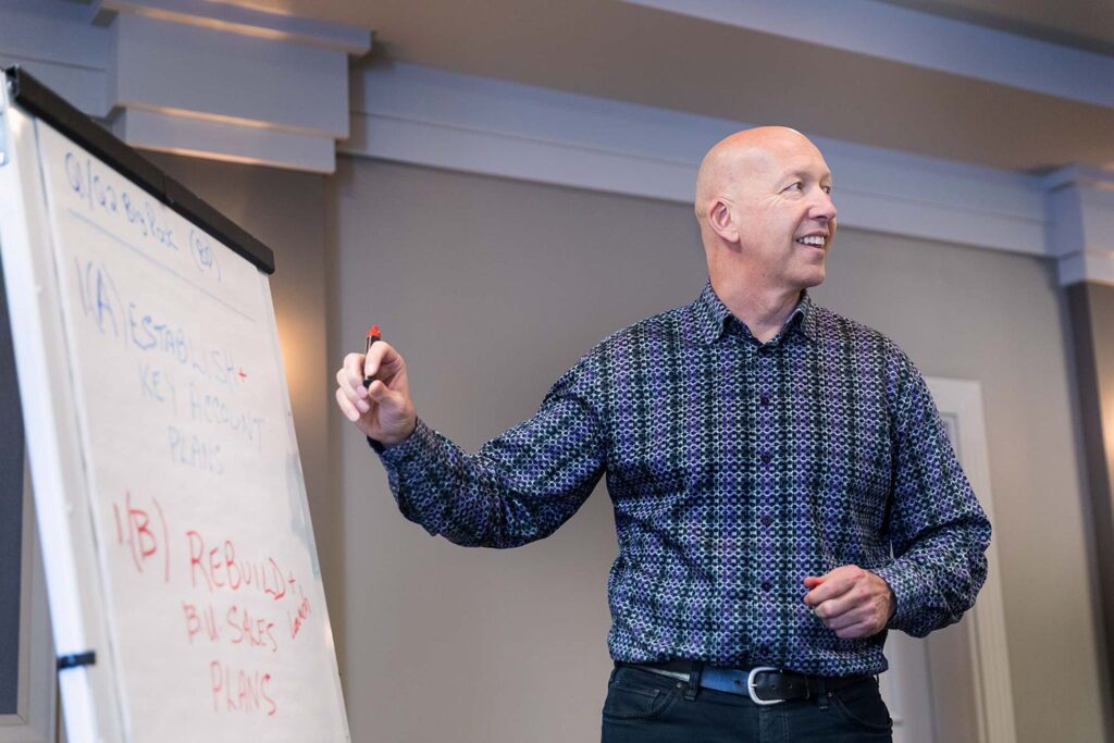 Professional leadership coaching session showing Ken Larson in patterned shirt leading a workshop or coaching session, gesturing towards a flip chart with written notes.