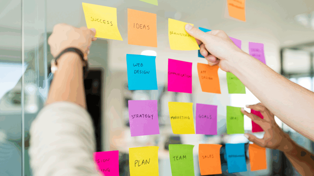 People arranging colorful sticky notes with ideas and project goals on a glass wall during a team planning session.