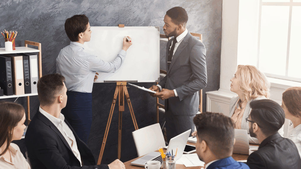 Business professionals participating in a collaborative team meeting, with one person writing on a whiteboard while others look on attentively.