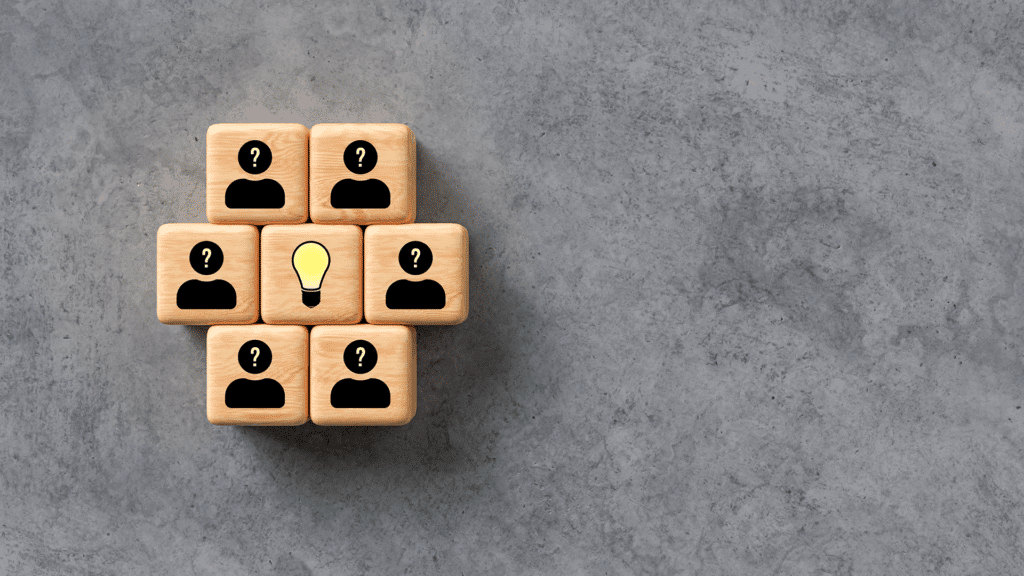 Wooden blocks with black silhouettes and question marks arranged around a central block featuring a yellow lightbulb, symbolizing teamwork and collective ideas.